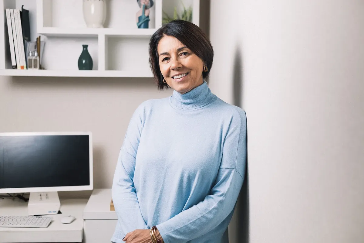 Rosane Vianna Gonzalez sorrindo, encostada na parede do consultório, ao lado da mesa e de uma estante branca