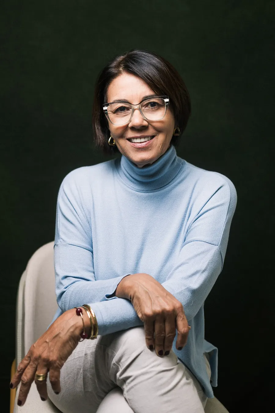 Rosane Vianna Gonzalez sorrindo, de óculos e blusa azul-clara, sentada diante de um fundo verde-escuro
